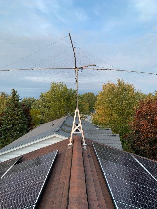 Radio antenna mounted on a white support structure on a rooftop with solar panels. Trees and blue sky background.