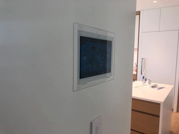 Tablet mounted in a white wall, displaying a blue screen. A light switch is below.