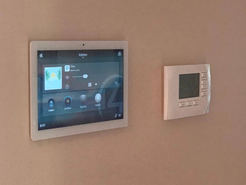 White tablet and thermostat mounted on a tan wall. Tablet displays music interface.