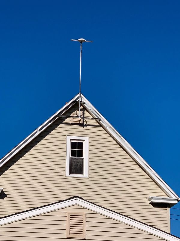 Tan house gable with white trim, small window, and tall antenna against a clear blue sky.