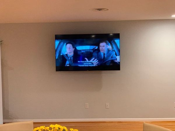 Wall-mounted TV showing a movie scene with two men in a car. Light-colored walls, minimalist decor.