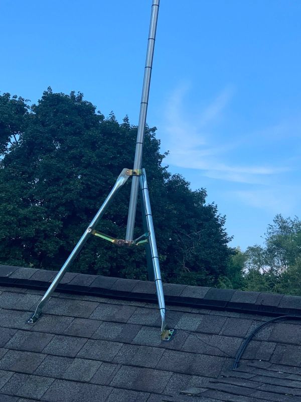 A gray radio antenna on a tripod mount is secured to a dark roof against a cloudy blue sky.
