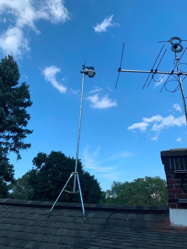 A camera on a tripod atop a roof, with a TV antenna and blue sky in the background.