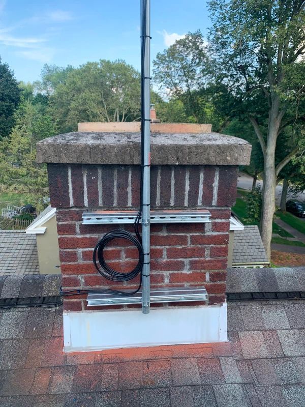 A chimney with a metal pole and cables attached to it. It sits on a roof with visible shingles, set against a background of trees.
