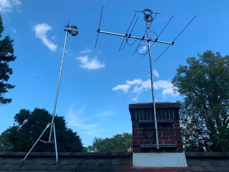 Two antennas on a rooftop against a blue sky with trees. One antenna is on a tripod; the other is mounted on a chimney.