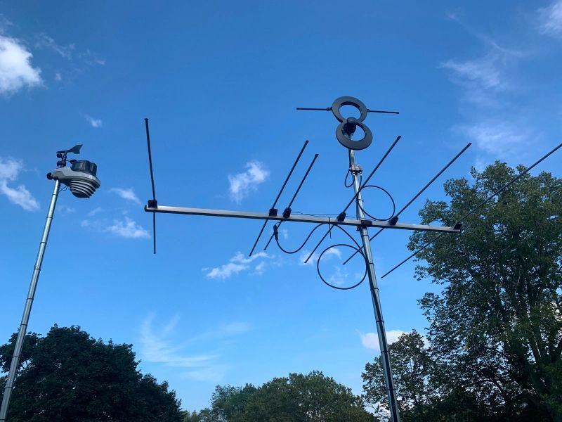Antenna array with circular and linear elements mounted on poles, against a blue sky with trees.