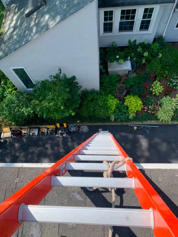 Orange ladder angled down from a roof. Tools sit on a driveway below a house with a garden.