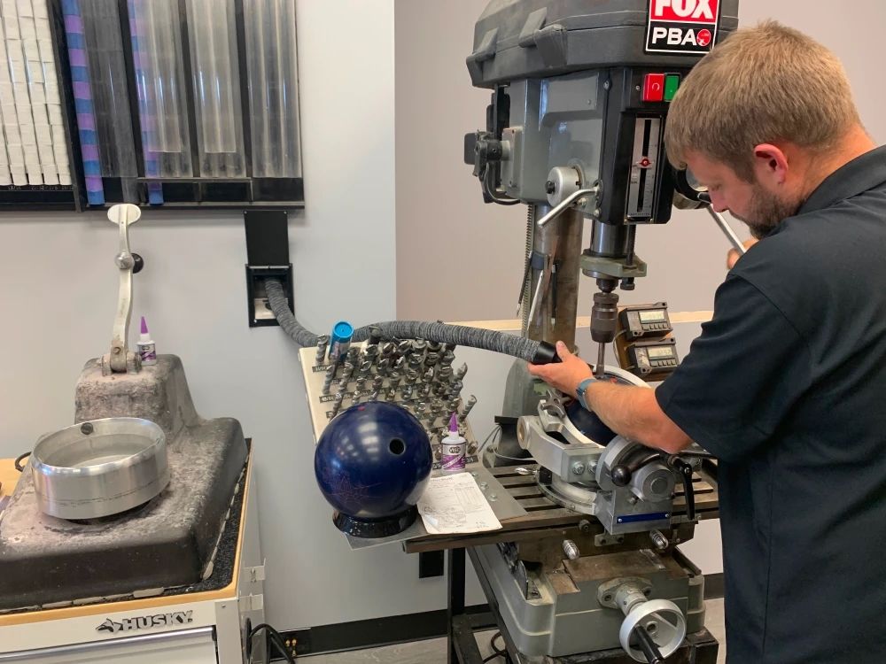 Man drilling a blue bowling ball with a drill press, in a bowling alley setting.