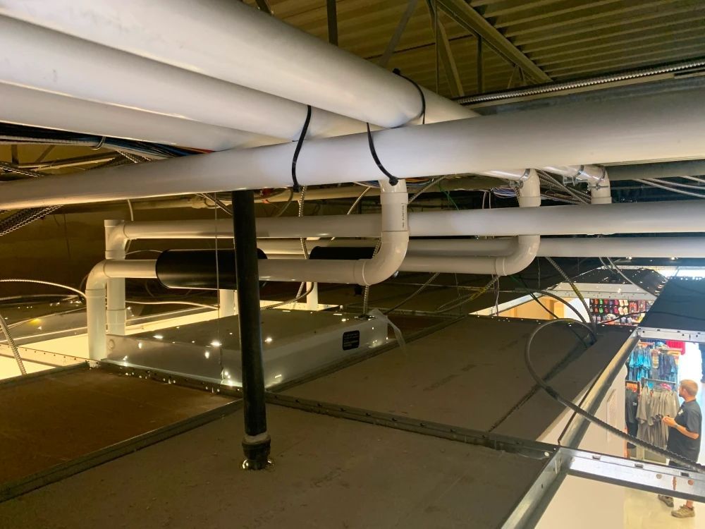 Pipes and ventilation ductwork attached to a ceiling with a metal structure below. People are visible in the background.