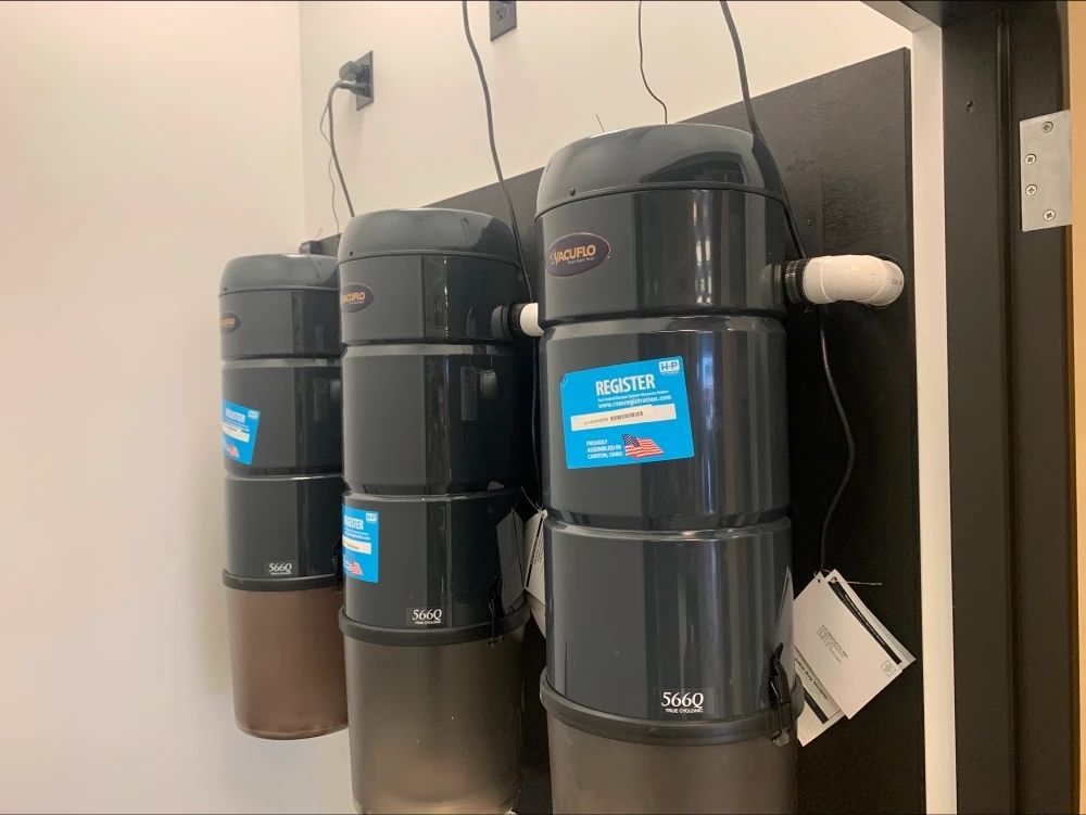 Three central vacuum units mounted on a wall. Dark gray cylinders with dust collection containers.