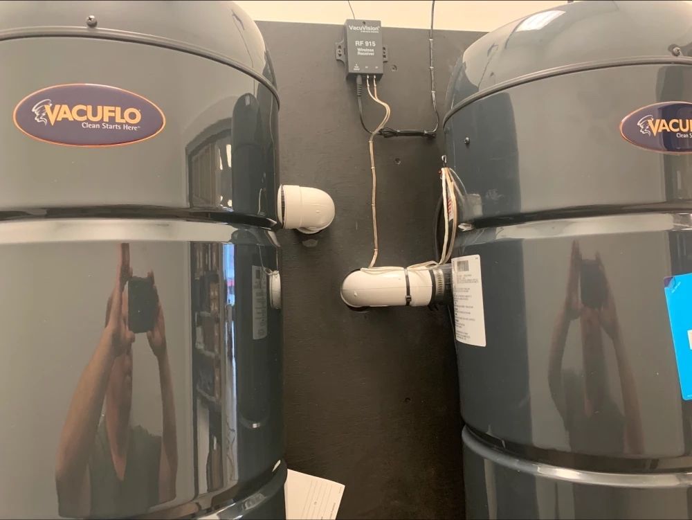 Two Vacuflo central vacuum units mounted on a dark wall. A person is reflected in the units, taking a photo.