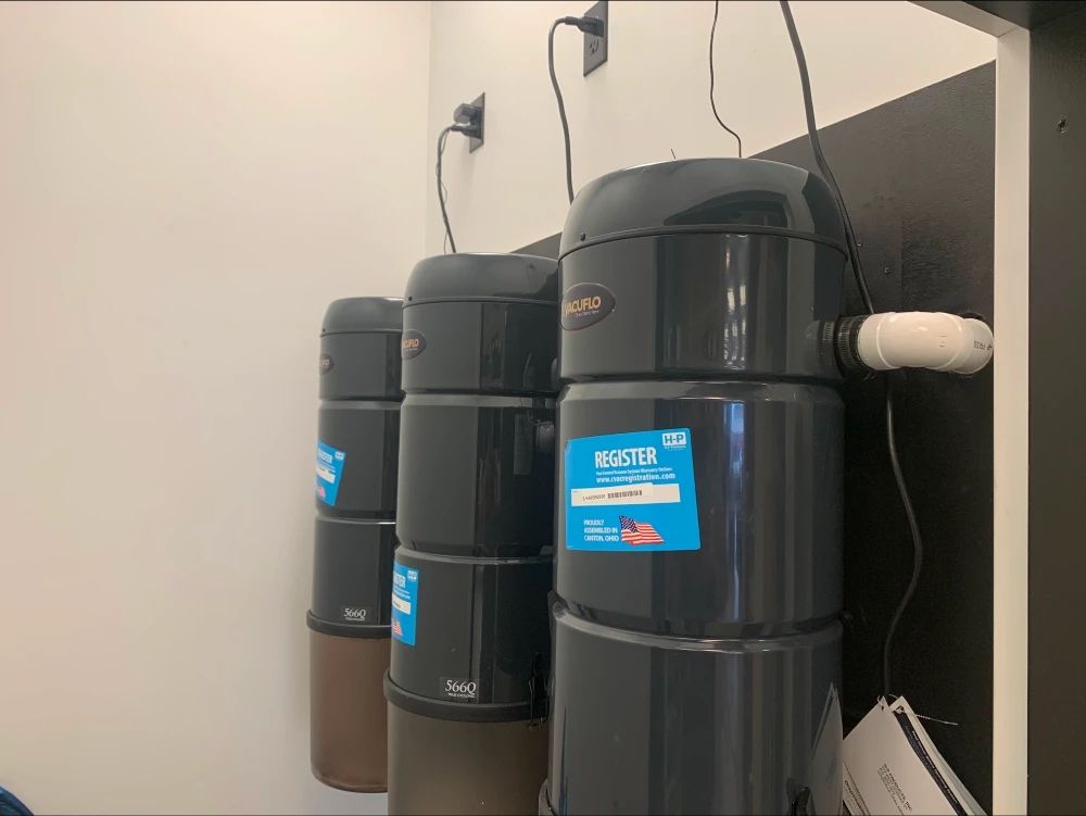 Three dark-colored central vacuum units mounted on a white and black wall, with power cords plugged in.