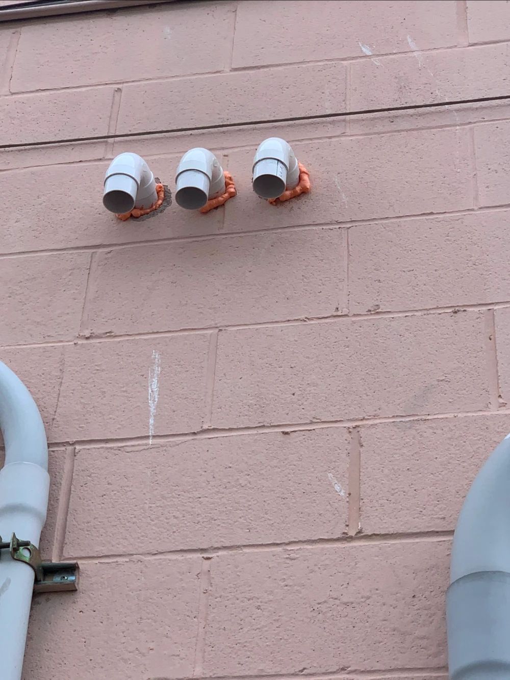 Three white vent pipes on a pink brick wall, held by orange supports.