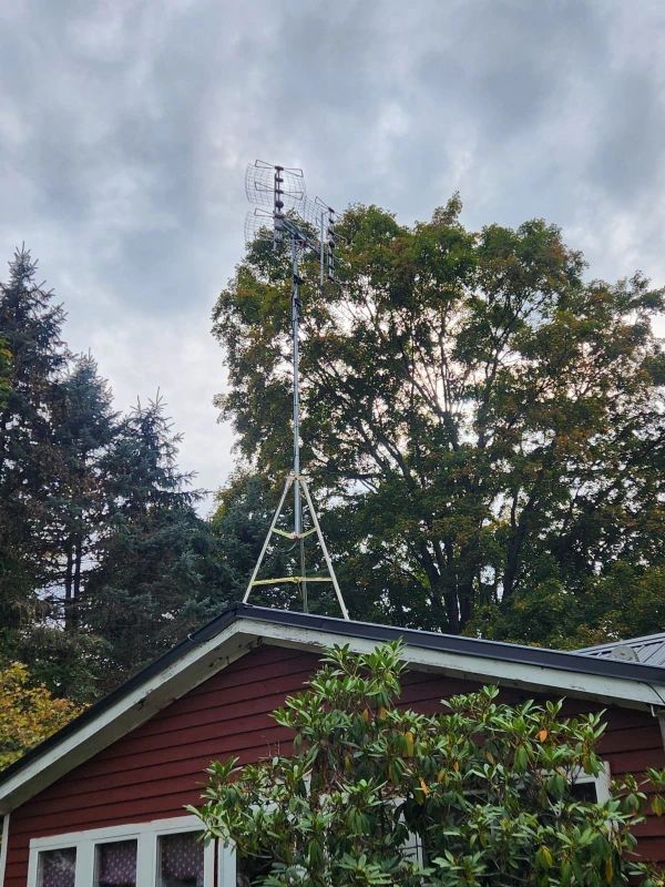 A tall metal antenna structure sits atop a red house, framed by trees and a cloudy sky.