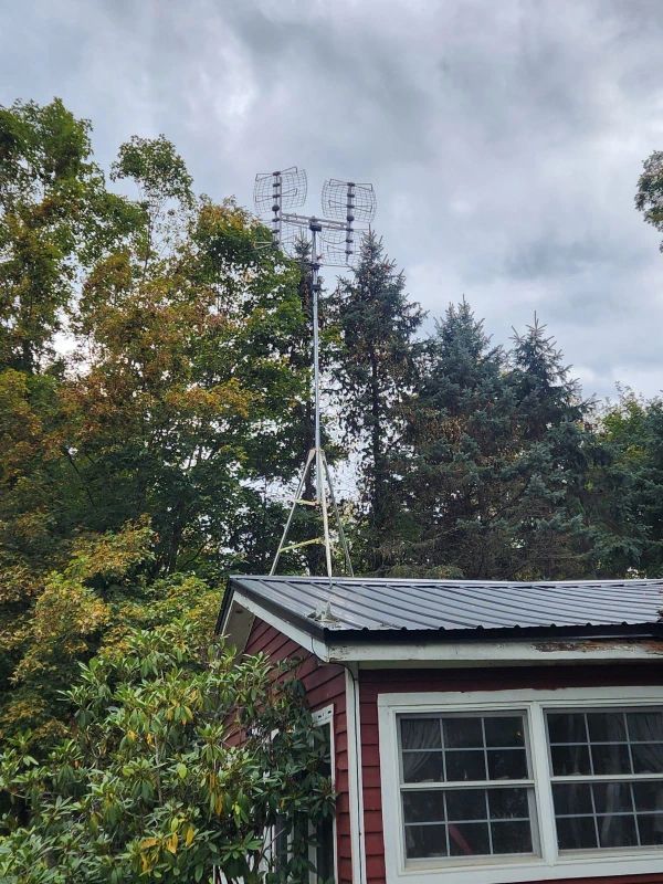 A tall antenna on a tripod above a red-roofed building, surrounded by trees under a cloudy sky.