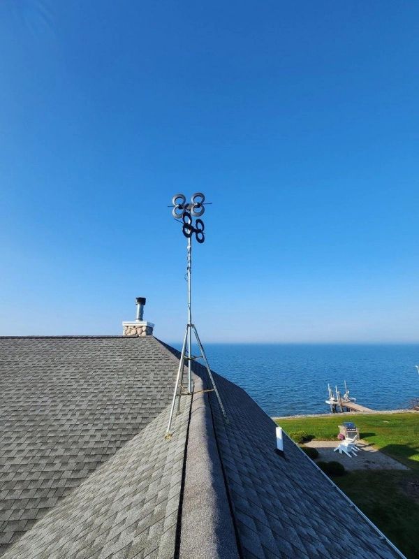 Anemometer atop a tripod on a dark shingle roof overlooking a body of water under a clear blue sky.