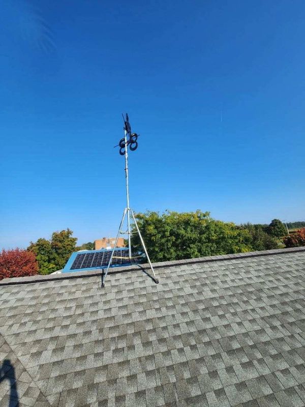 Solar panel and weather station equipment on a rooftop, set against a bright blue sky.