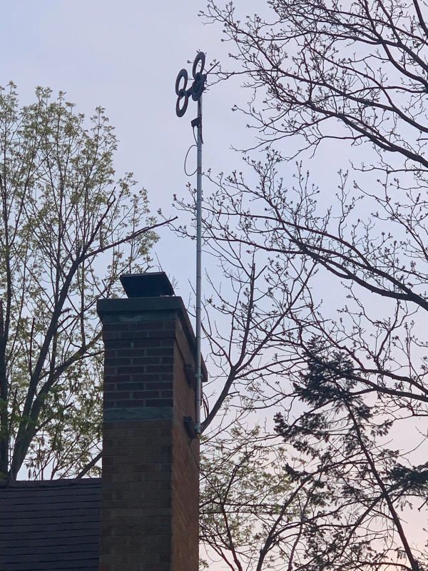 Chimney with a tall, thin antenna topped by a circular device, trees in the background.