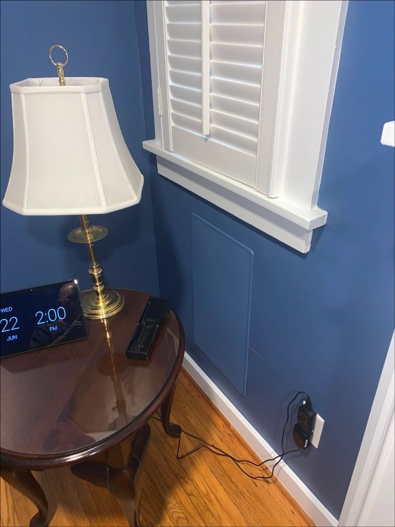 A nightstand with a lamp and electronics next to a blue wall with a window and an outlet.