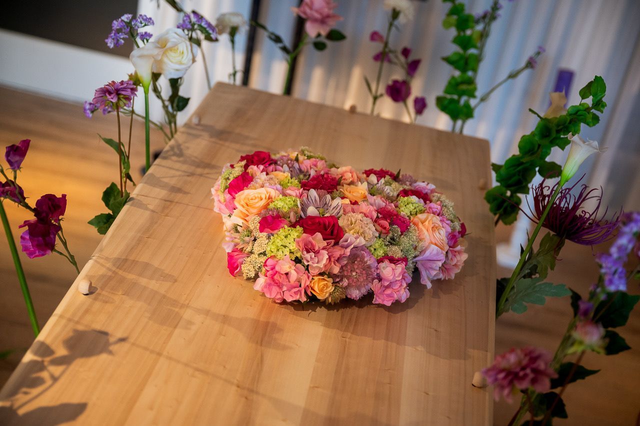 Ovaal rouwstuk van zijden bloemen in roze, rood en perzik op een lichte houten kist