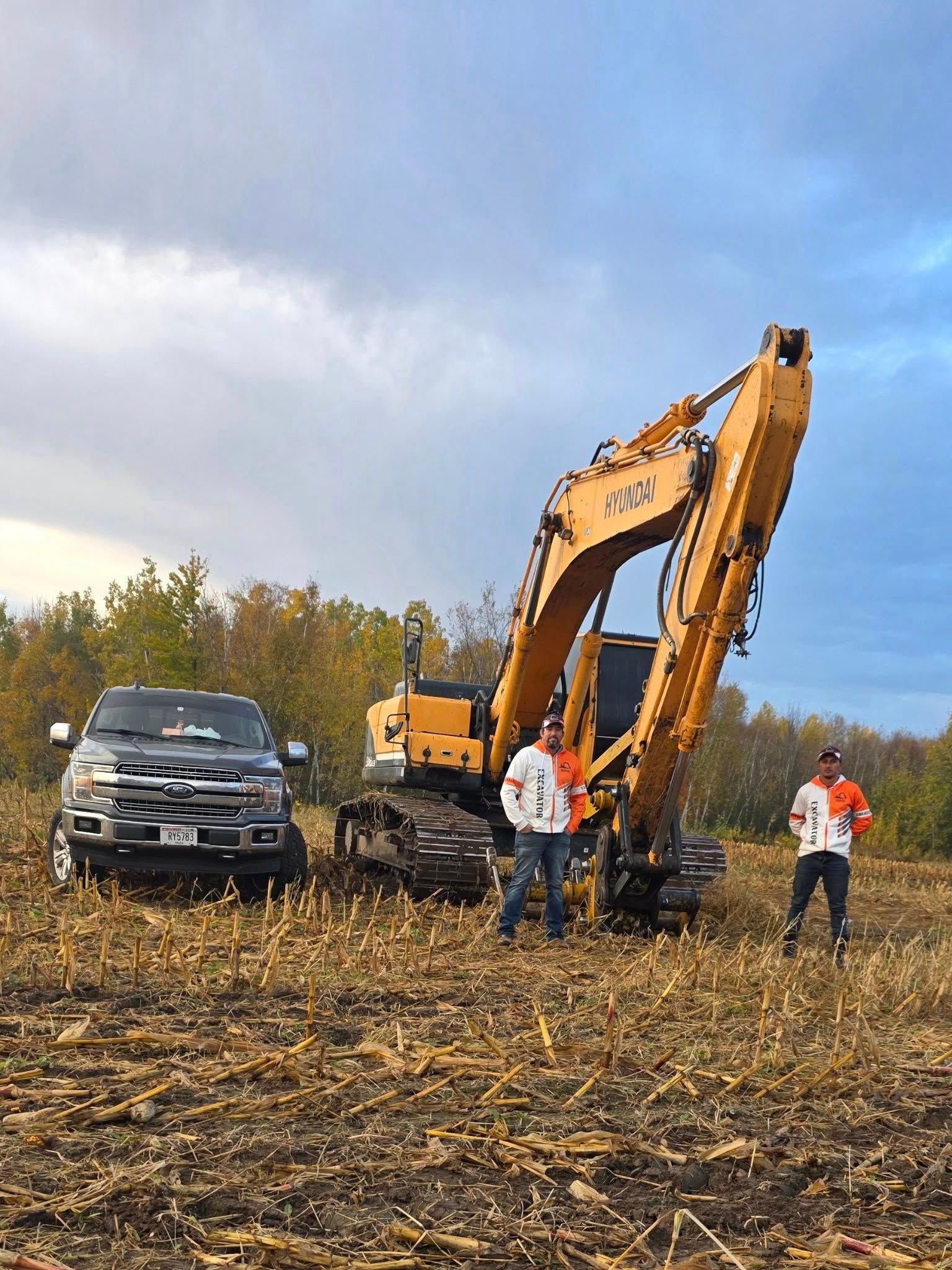 Two people stand in a harvested field next to a large yellow excavator and a dark pickup truck under a cloudy sky.