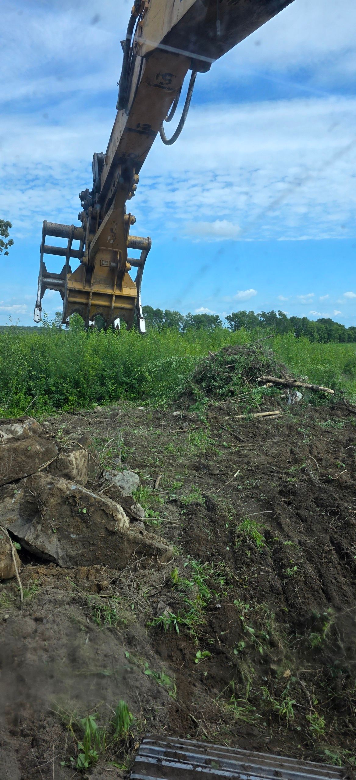 An excavator arm with a large mechanical grapple attachment extends high above a dirt pile in a grassy field.