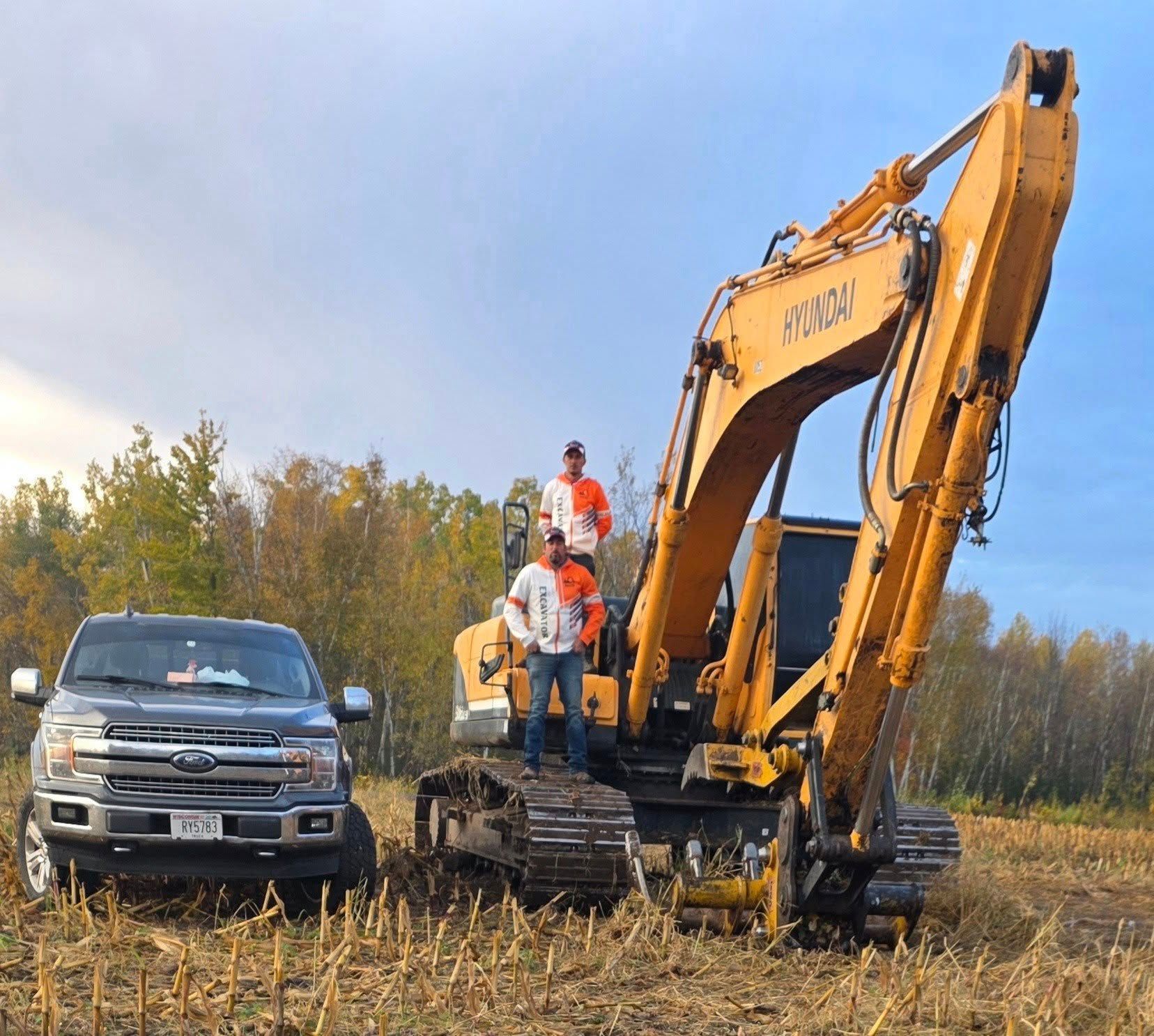 Two people stand on a large yellow excavator next to a grey pickup truck in a harvested cornfield under a blue sky.