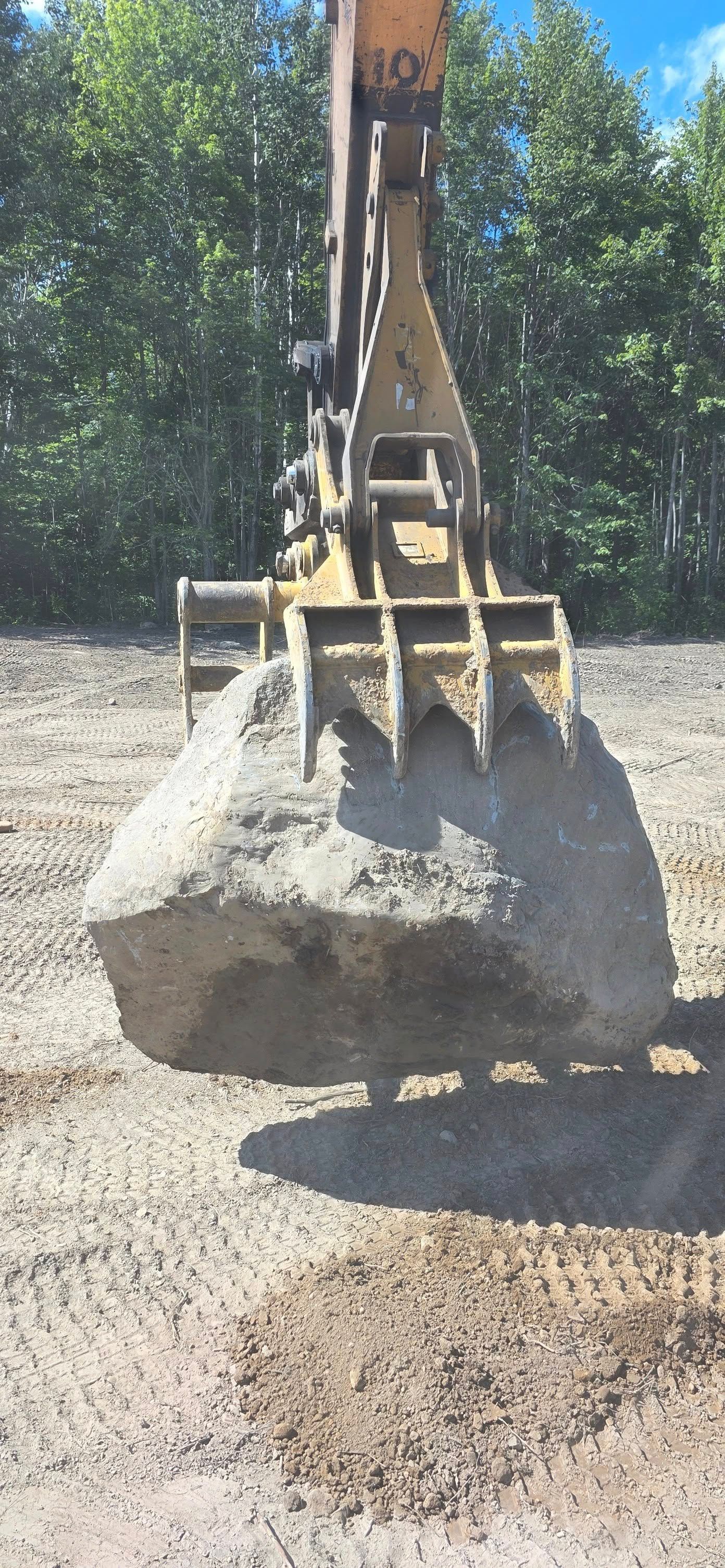 A large yellow excavator thumb attachment grips a massive gray boulder in an outdoor, sunny setting.