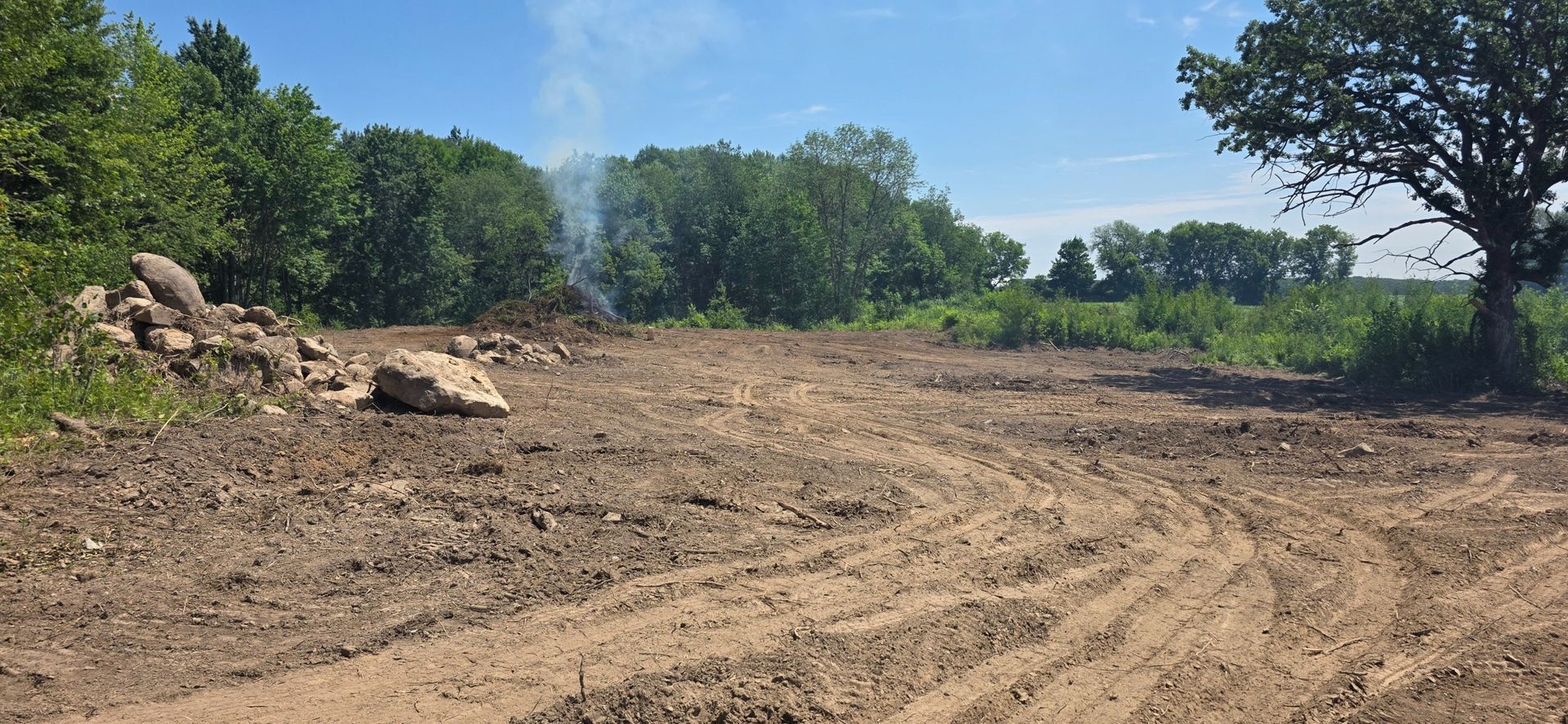 A field cleared of vegetation with piles of rocks, tire tracks in the dirt, and a line of trees against a clear blue sky.