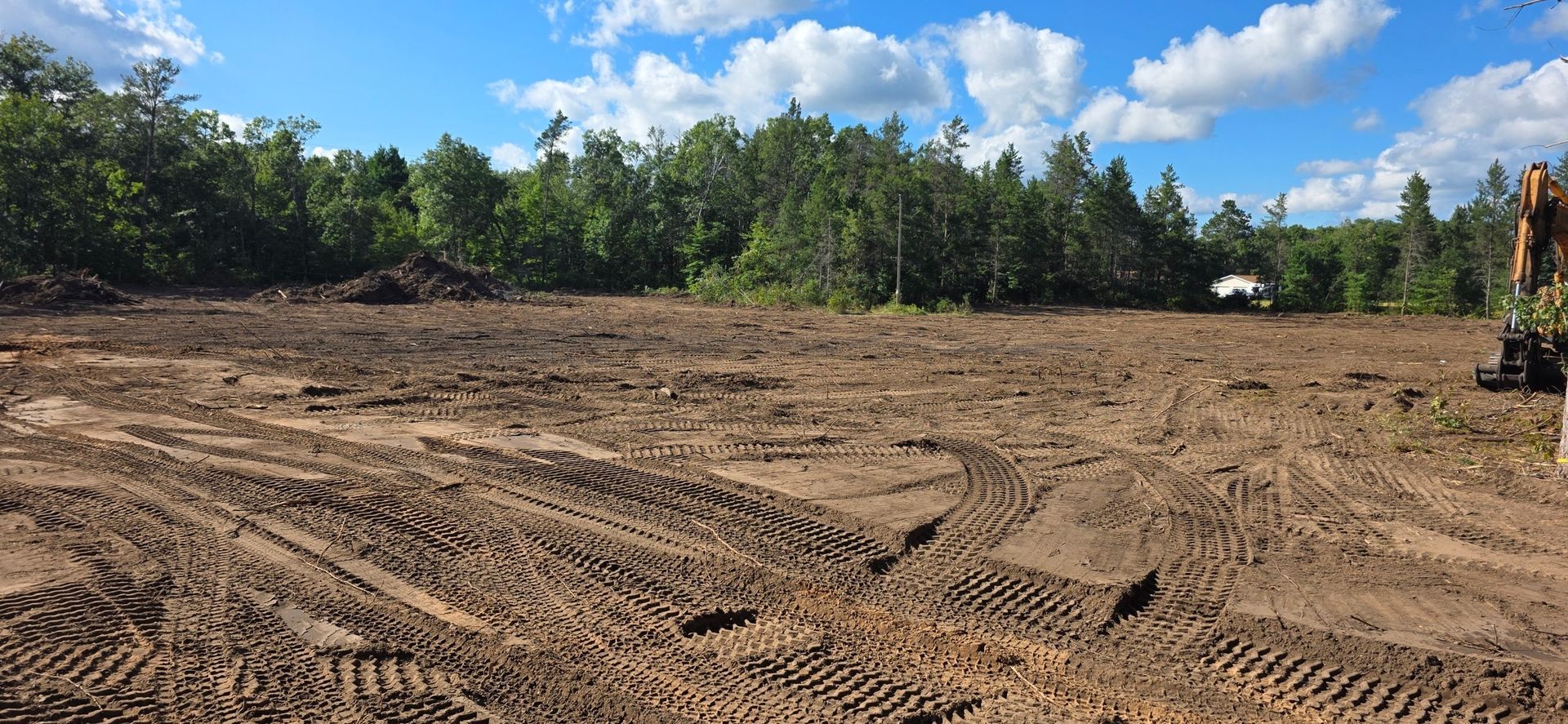 A cleared construction site with dirt tracks and tree lines under a blue sky with scattered clouds.