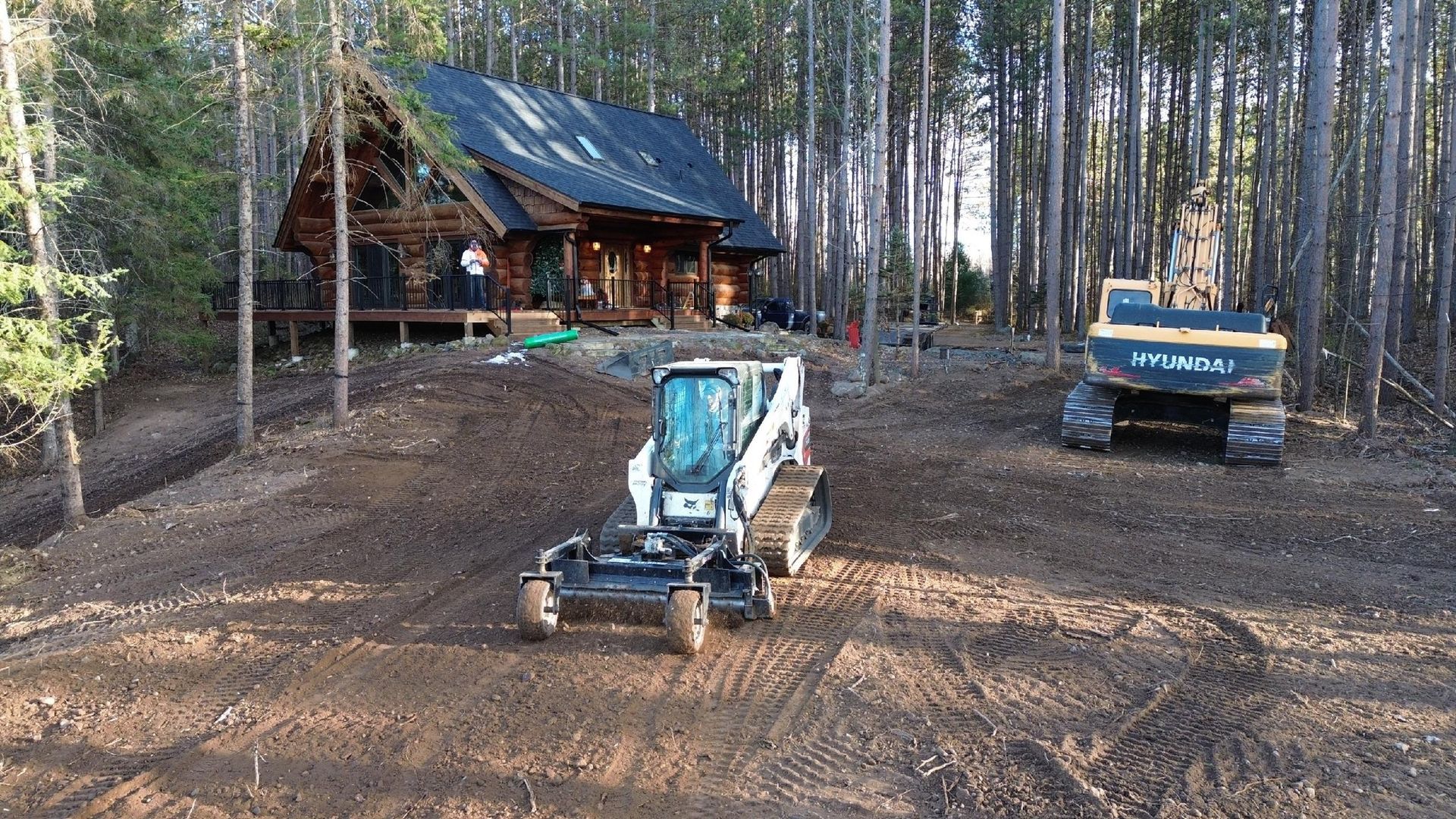 A track loader and an excavator sit on a dirt lot in a wooded area in front of a log cabin.