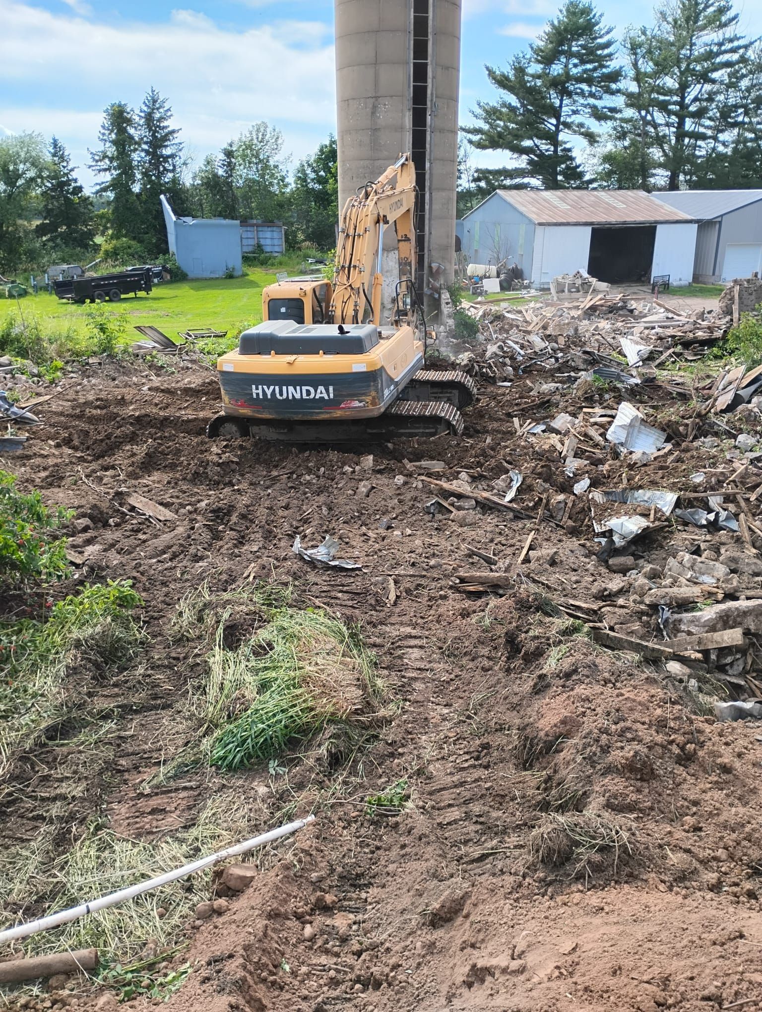 A yellow Hyundai excavator works on a construction site next to a tall concrete silo with debris scattered on the ground.