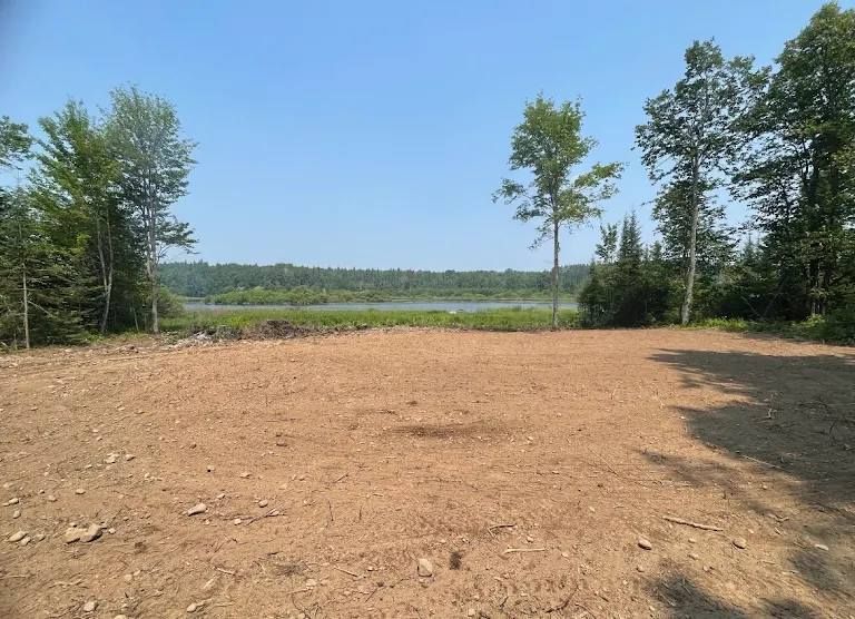 A clear, flat dirt lot surrounded by trees, with a view of a lake and a forested shoreline in the distance.