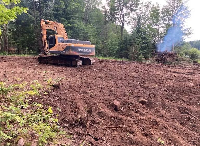 An orange excavator parked on cleared brown earth with a smoking brush pile in the background.