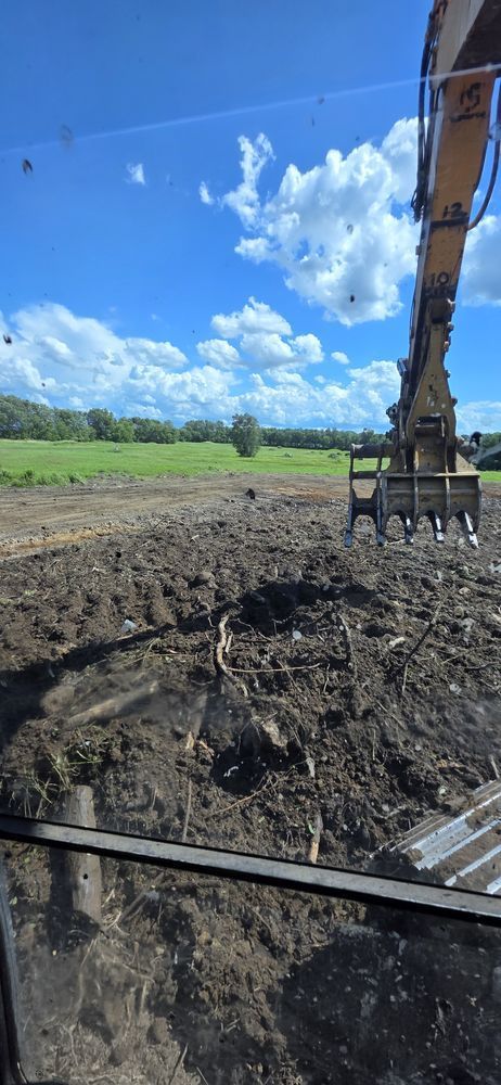 A view from a construction vehicle cab showing an excavator arm over tilled earth with a bright blue sky and fields.