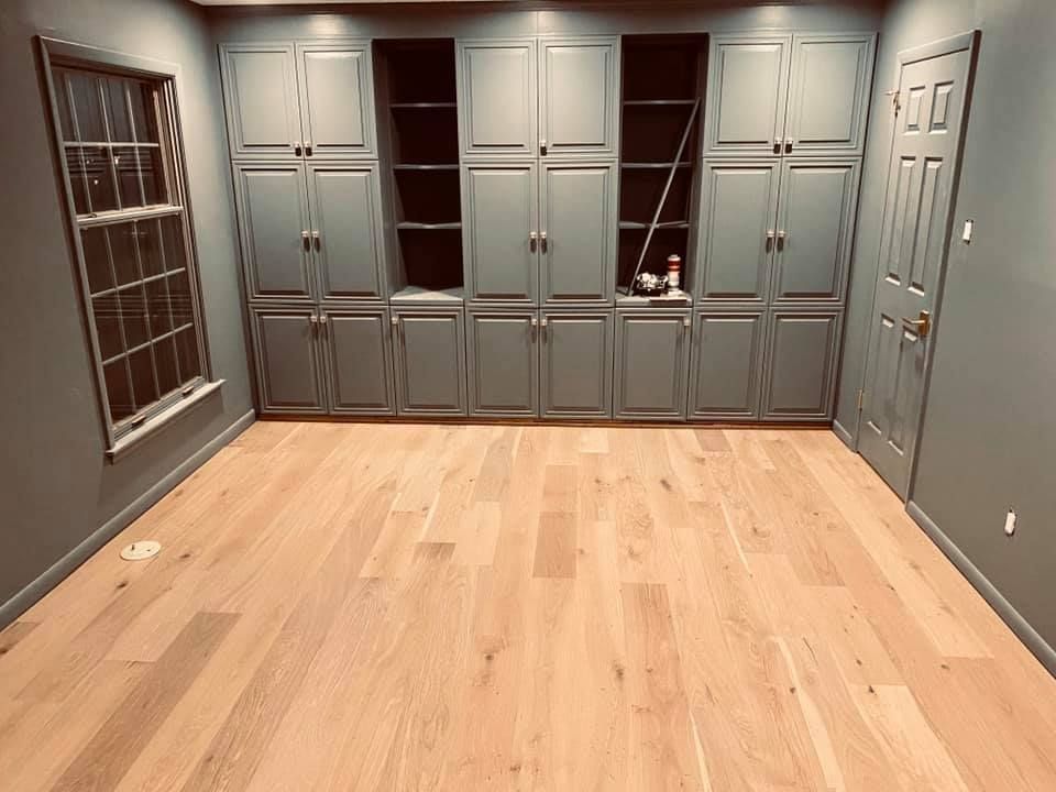 Empty room with light wood floors, gray walls, built-in cabinets, and windows on one side.