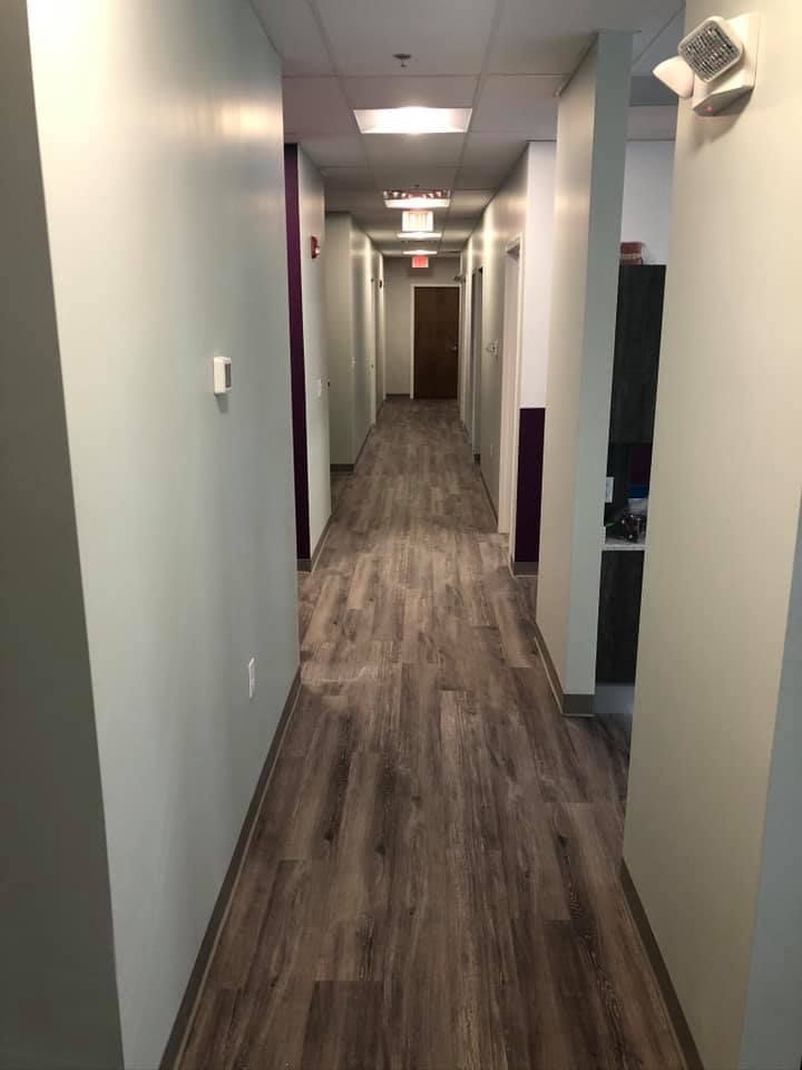Empty office hallway with gray walls, wood-look floor, and overhead fluorescent lights.