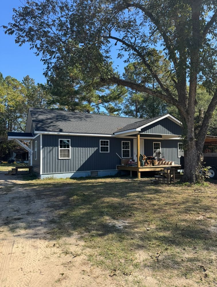 A single-story gray house with a dark roof and a small wooden front deck, surrounded by trees on a dirt lot.