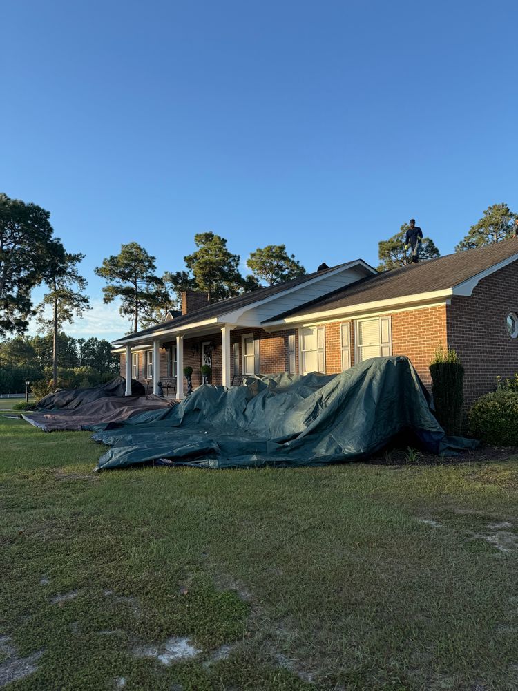 Workers replace a roof on a brick ranch house, with large protective tarps covering the front lawn.