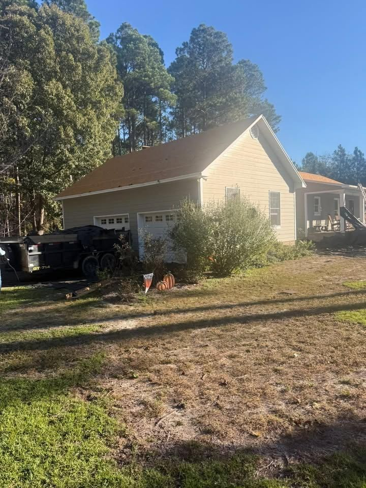 Small beige house with a black truck in the driveway, surrounded by trees and yard in bright sunlight