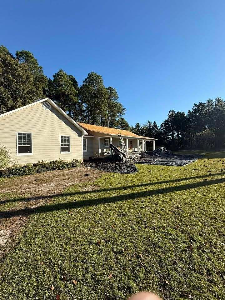 A light-colored house with a wooden porch roof under construction, surrounded by a lawn and trees under a clear blue sky.