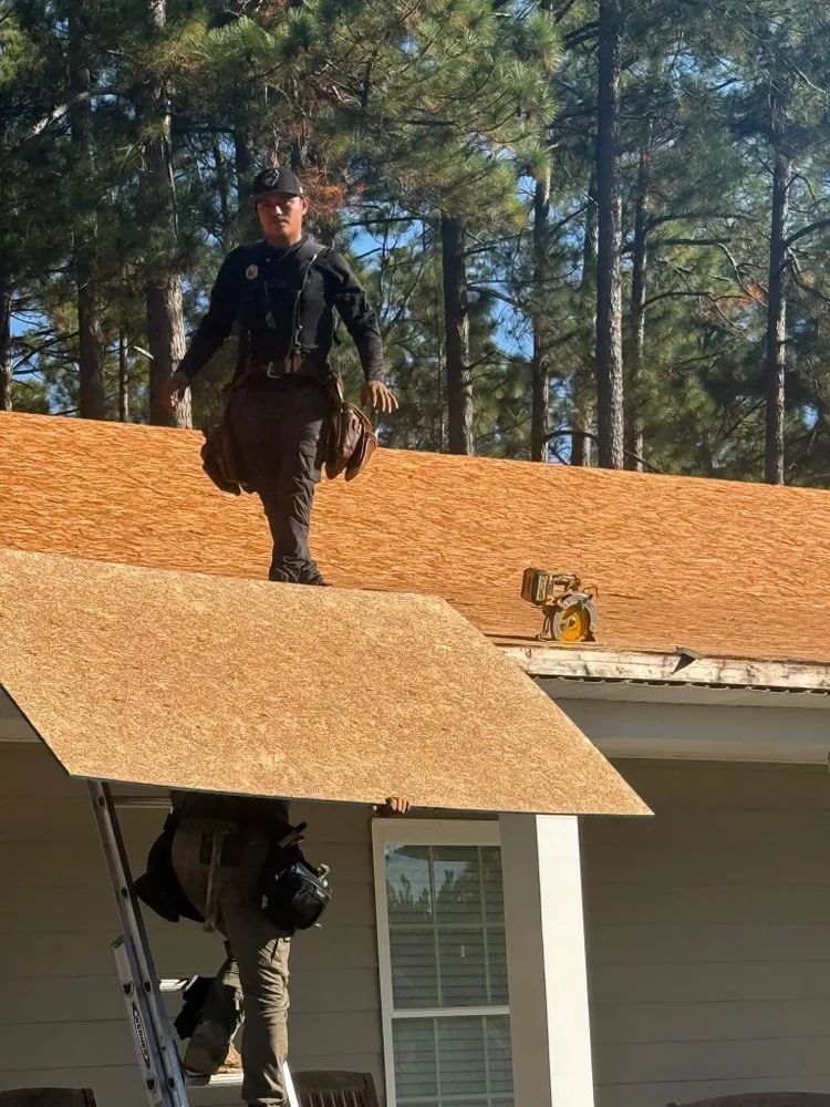 A worker in dark clothing stands on a plywood-covered roof next to a ladder during a home renovation.