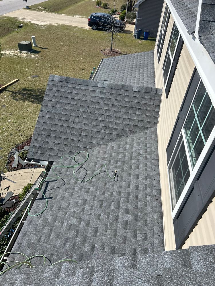 An elevated, angled view of a gray asphalt shingle roof on a house with beige siding and a window, overlooking a yard.