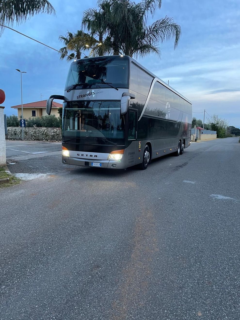 Un grande autobus a due piani color argento è parcheggiato su una strada asfaltata. È il crepuscolo e la scena è all'aperto.