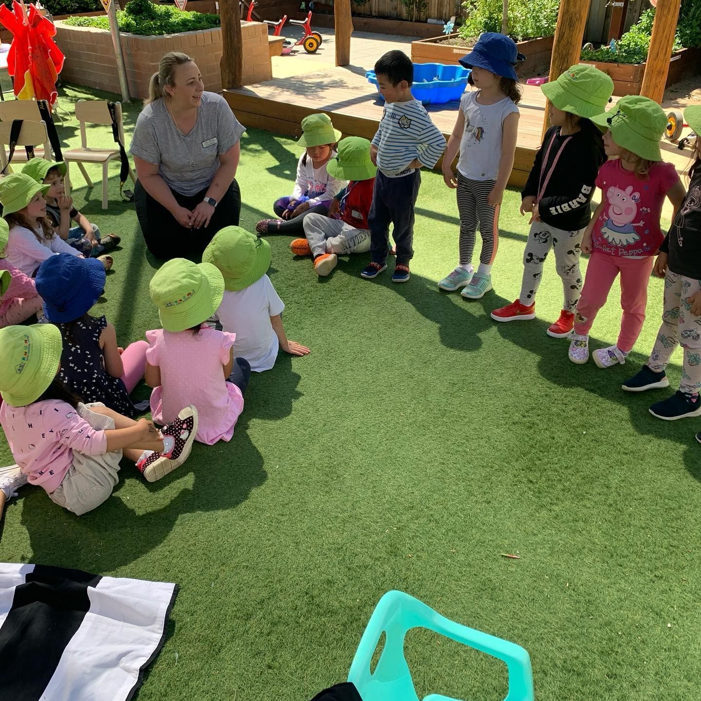 A group of children in hats sit in a circle on artificial grass with an adult for an outdoor preschool activity.