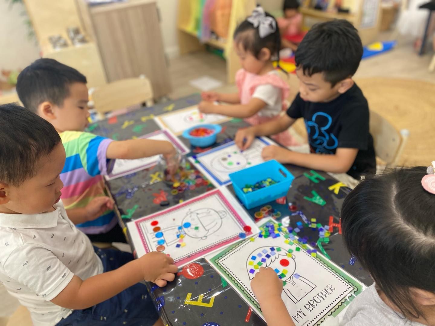 Four children sit at a table in a classroom, focused on placing colorful beads onto printed activity sheets.