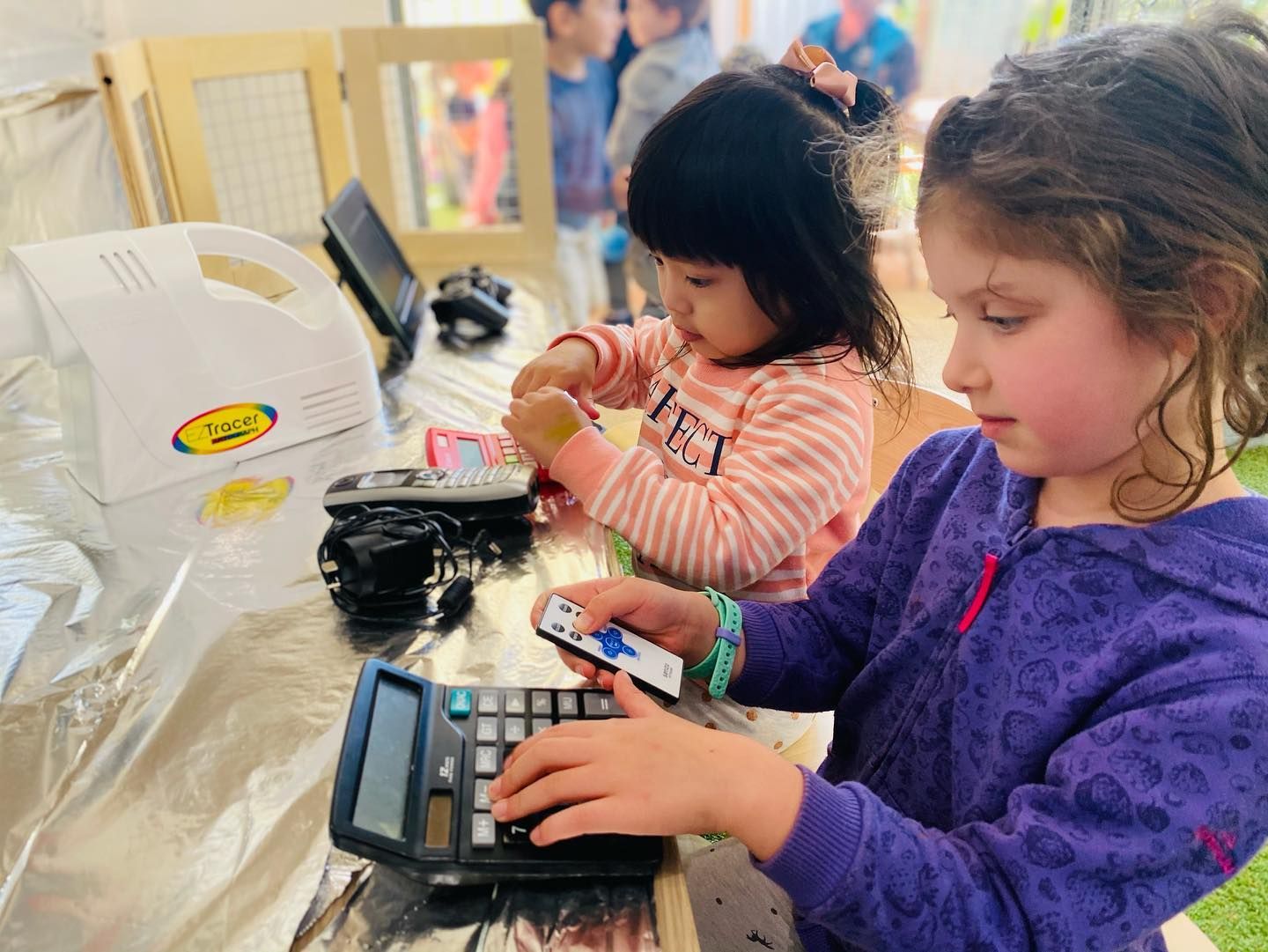 Two children sit at a table playing with calculators and electronic devices in a classroom setting.