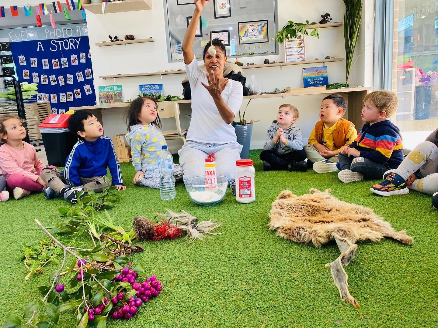 An educator leads an indoor circle time with six children on artificial grass, demonstrating with white powder and plants.