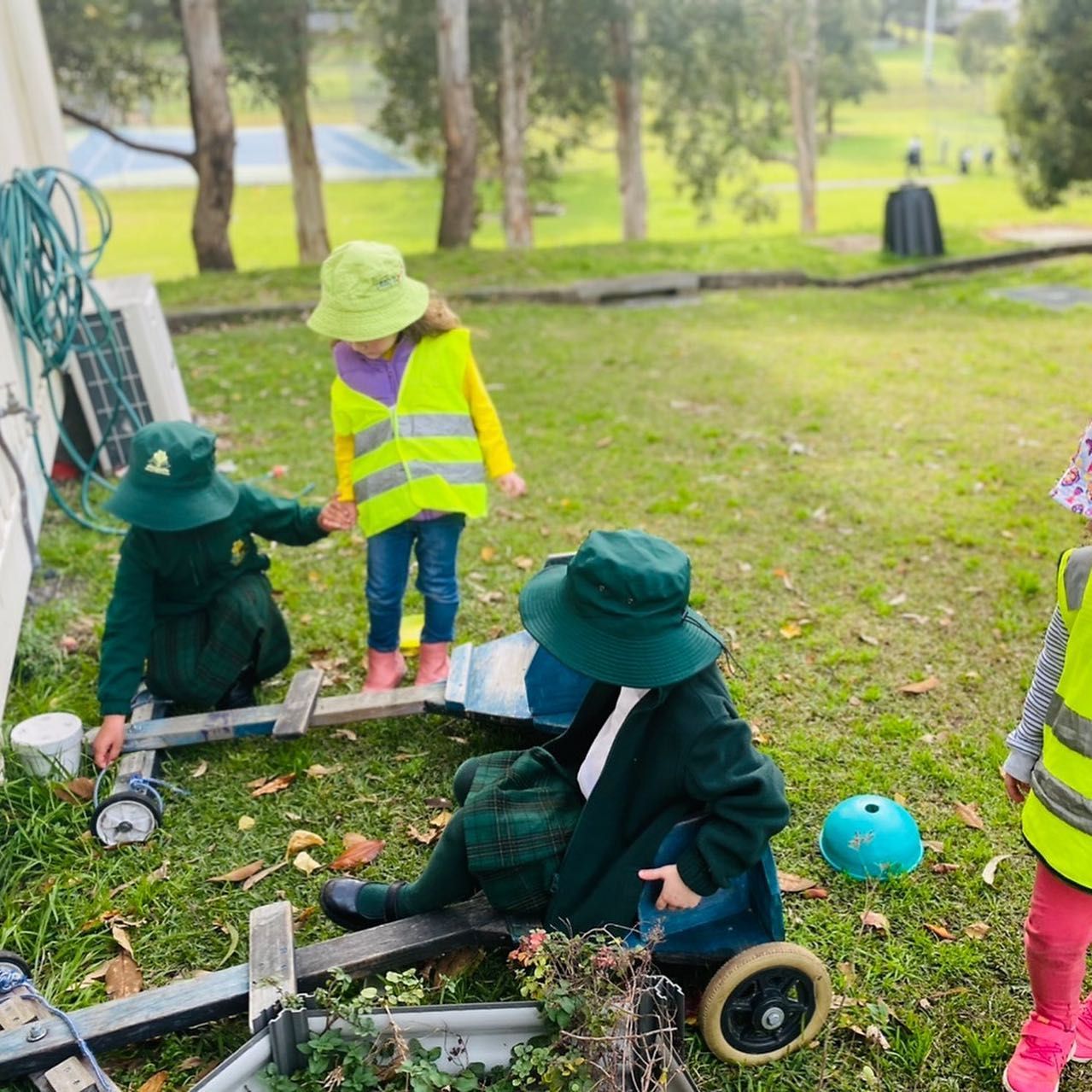 Four children in school uniforms and high-visibility vests play with a wooden toy vehicle on a grassy outdoor area.