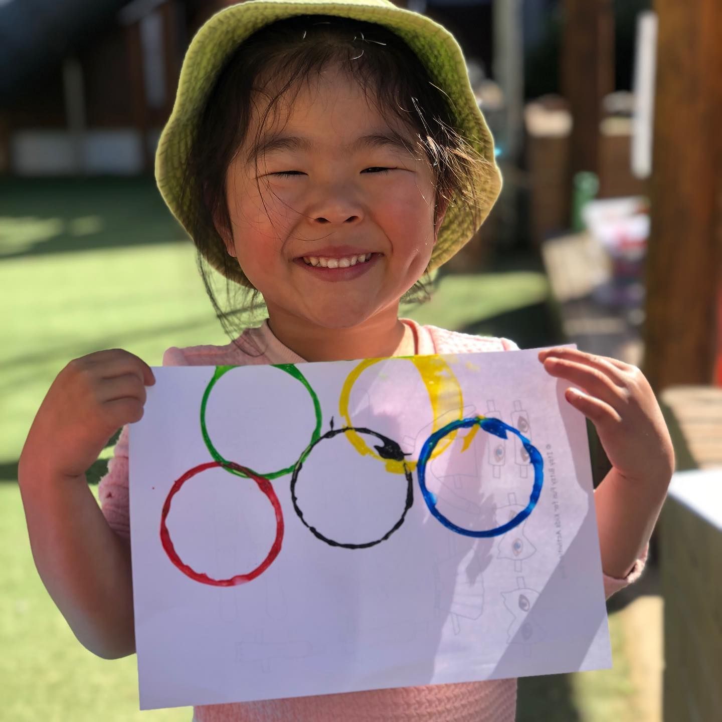 A smiling child in a green hat holds a piece of paper painted with five colorful circles arranged like Olympic rings.